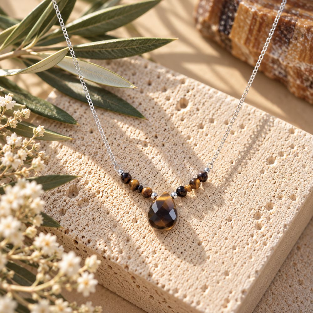 Tiger’s Eye beaded necklace featuring a faceted brown and golden teardrop pendant on a delicate silver chain, displayed on natural beige stone with olive branches and white flowers.