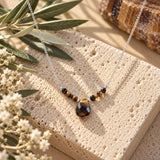 Tiger’s Eye beaded necklace featuring a faceted brown and golden teardrop pendant on a delicate silver chain, displayed on natural beige stone with olive branches and white flowers.
