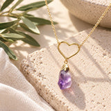 Gold open-heart necklace with a dangling raw purple amethyst crystal pendant, displayed on textured natural stone with soft greenery and ivory linen in natural daylight.