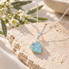 Raw aquamarine crystal necklace with a silver chain and prong-set pale blue gemstone pendant, displayed on textured natural stone with delicate white flowers.