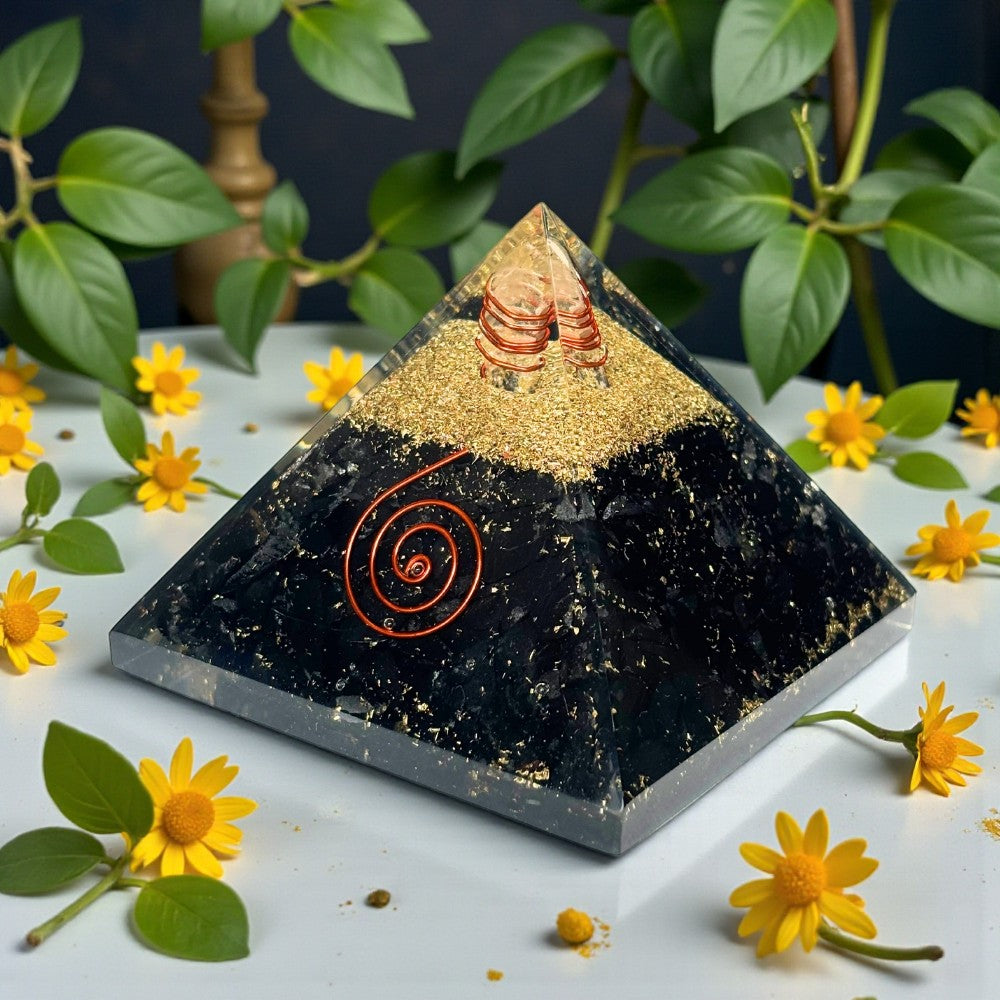 Black pyramid crystal with gold and red accents on a white surface with yellow flowers and green leaves.