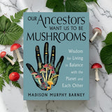 The book 'Our Ancestors Want Us to Be Mushrooms' by Madison Murphy Barney is displayed flat on a white marble surface, surrounded by green fern leaves and small red-and-white mushrooms. The book cover is light blue with black text and a central illustration of a hand made of mushrooms.