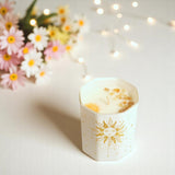 Candle in a decorative holder with sun design, surrounded by flowers and string lights.