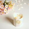Candle in a decorative holder with sun design, surrounded by flowers and string lights.
