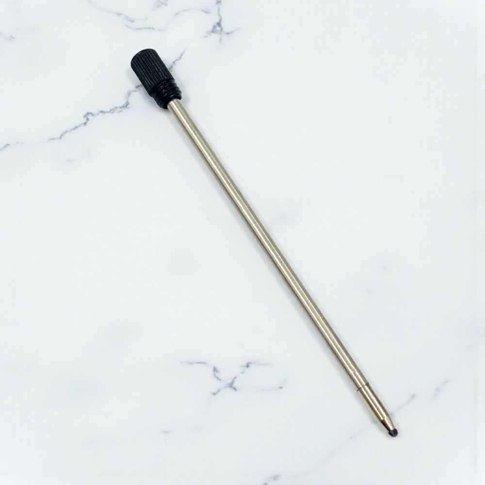A replacement pen refill with a long metallic silver body and a black textured cap, placed diagonally on a white marble-patterned background.