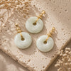 Jade Donut Pendant with natural green and white jade stone donuts arranged on a textured beige stone surface with soft linen fabric and dried flowers.