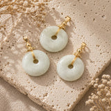 Jade Donut Pendant with natural green and white jade stone donuts arranged on a textured beige stone surface with soft linen fabric and dried flowers.