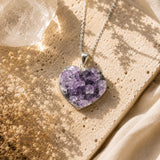 Heart-shaped purple crystal pendant on a chain, placed on a textured surface with a natural light background.