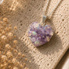 Heart-shaped purple druzy pendant on a silver chain against a textured beige background with dried flowers.