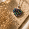 Heart-shaped pendant necklace with a multicolored stone on a textured surface with dried flowers.