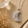Crescent moon-shaped pendant necklace on a textured beige surface with dried flowers.