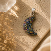 Multicolored crescent moon pendant on a textured beige surface with dried flowers.