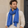 Man wearing a blue scarf with pom-pom details, standing against a beige wall.