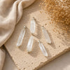 Five clear crystal point pendants with silver-toned caps arranged on a beige stone slab with a beige fabric and dried flowers in the background.