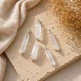 Five clear crystal point pendants with silver-toned caps arranged on a beige stone slab with a beige fabric and dried flowers in the background.