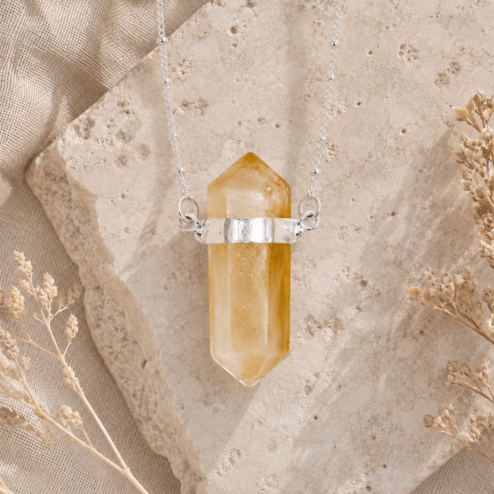 A silver pendant necklace featuring a faceted yellow crystal point hanging from a delicate silver chain, styled on a textured beige stone slab with dried flowers.