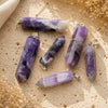 Five purple and white amethyst crystal point pendants with silver bail loops, arranged on a textured beige stone surface with dried floral props.