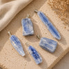 Five blue-and-white striped stone pendants with silver bail loops, arranged on a textured beige stone surface with a beige fabric and dried flowers in the background.