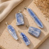 Five blue-and-white striped stone pendants with silver bail loops, arranged on a textured beige stone surface with a beige fabric and dried flowers in the background.
