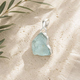 Raw aquamarine pendant with a sterling silver wrap resting on a textured natural stone surface with soft linen accents and botanical greenery.