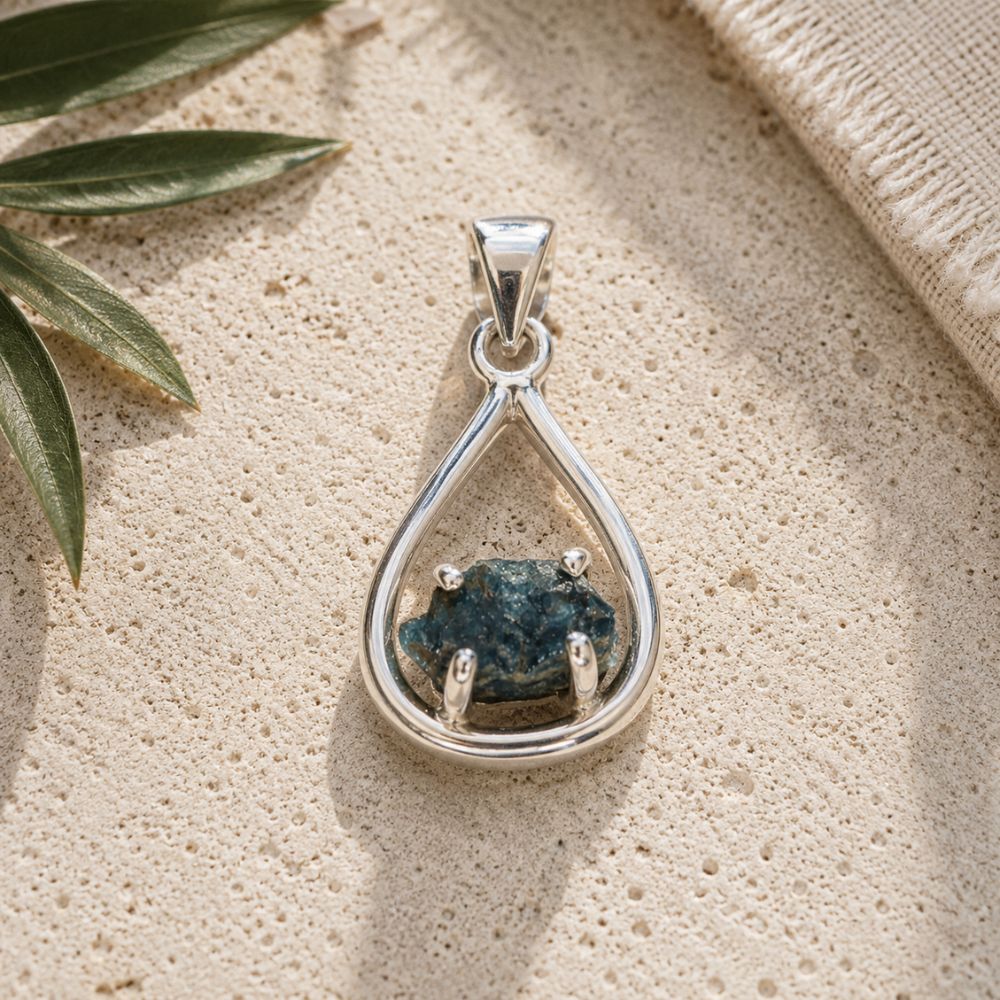 Raw Blue Apatite silver teardrop pendant displayed on a natural stone surface with soft daylight. Each pendant features a unique raw blue apatite crystal in a polished silver teardrop setting.