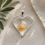 Raw citrine heart pendant featuring a natural citrine crystal suspended inside an open silver heart frame, photographed on a neutral travertine surface in soft natural light.