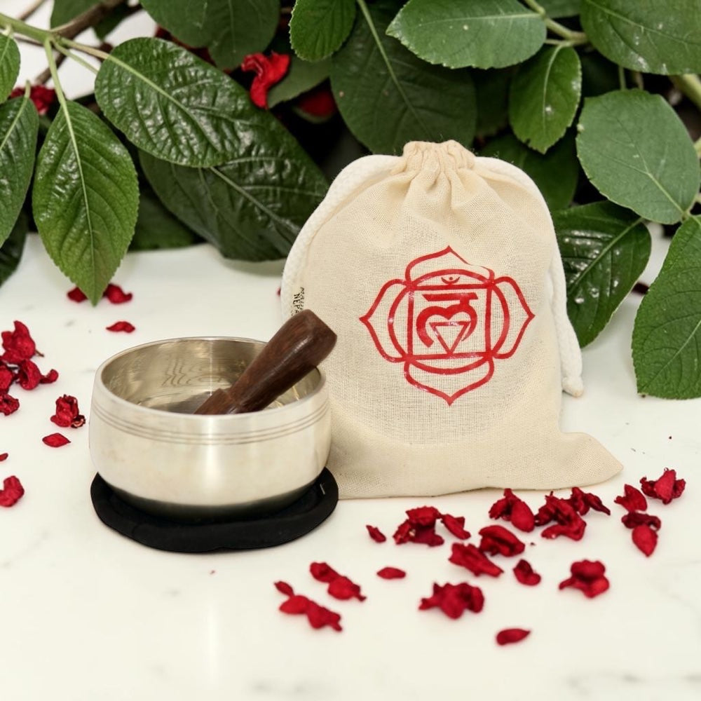 Meditation set with metal bowl, wooden stick, and drawstring bag with rose emblem on a white surface with red flowers and green leaves.