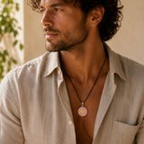 A man wearing a light beige linen shirt and a black cord necklace with a round pink stone pendant, shown in a warm, softly lit setting.