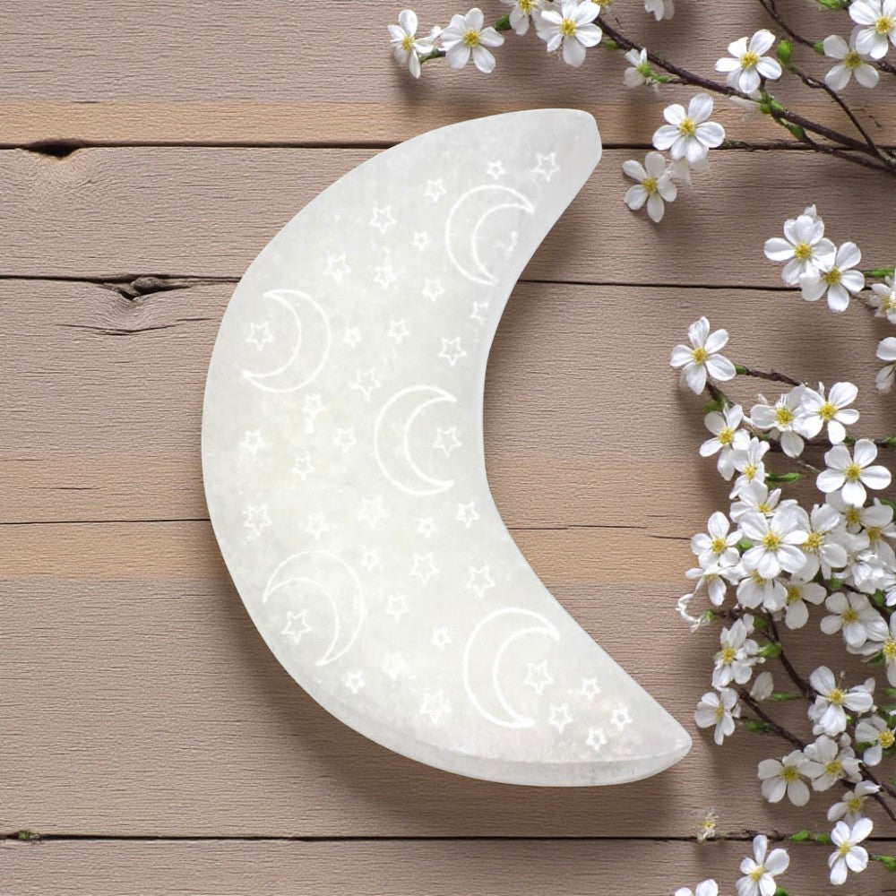 Decorative moon-shaped selenite charging plate with floral design on a wooden surface with white flowers.