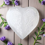 Heart-shaped selenite with engraved angel wings on a wooden surface with purple flowers.