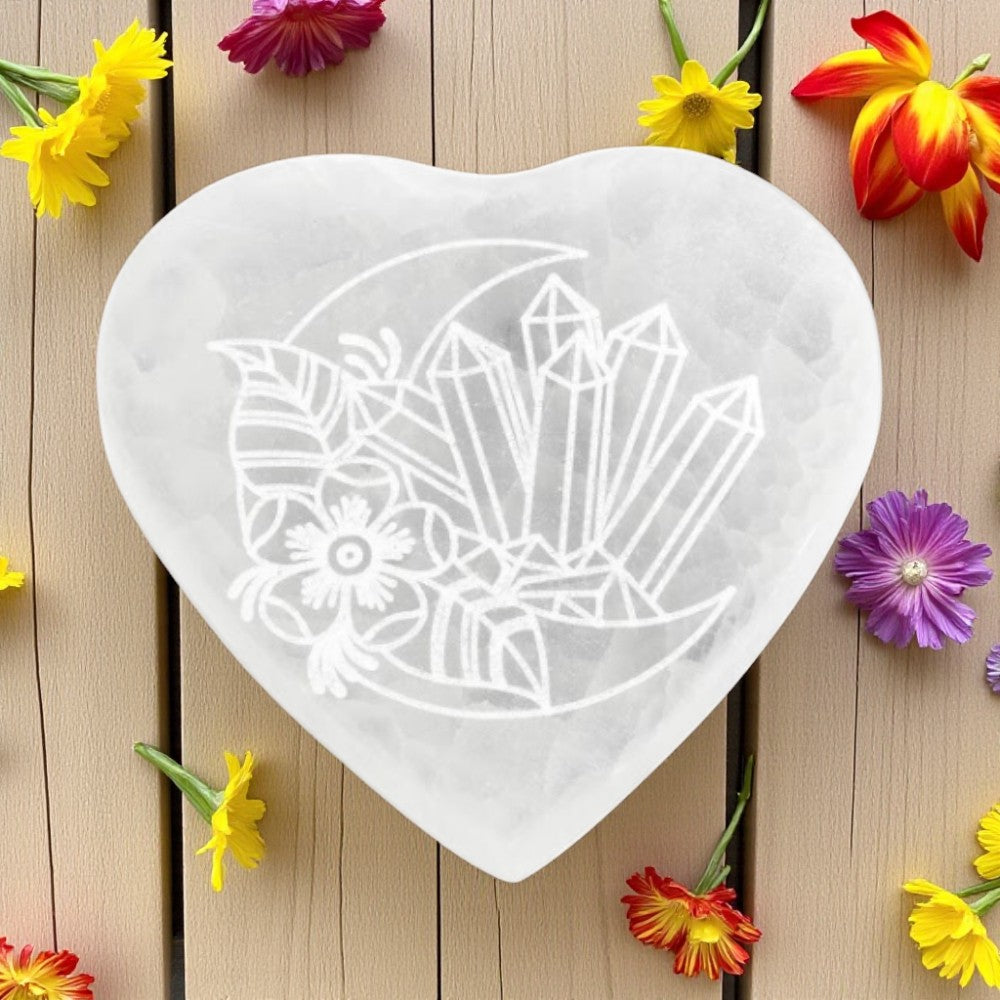 Heart-shaped selenite charging plate with stenciled design on a wooden surface with colorful flowers.
