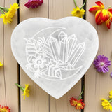Heart-shaped selenite charging plate with stenciled design on a wooden surface with colorful flowers.