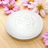 White selenite charging plate with intricate design on a wooden surface with pink flowers.