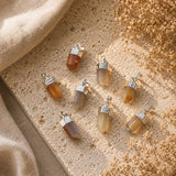 Six small pendant charms with translucent stone drops and silver caps, arranged on a textured beige surface with linen and dried flowers.