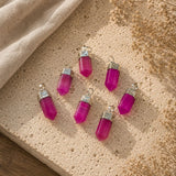 Five magenta-pink crystal pendants with silver caps and loops, arranged on a textured beige stone slab with a linen cloth and dried flowers.