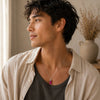 A man wearing a silver-toned pendant necklace with a vibrant magenta-pink rectangular stone, styled with a beige linen shirt over a dark gray t-shirt in a softly lit indoor setting.