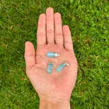 Three small light blue crystal point pendants with silver jump rings are held in the palm of a hand, set against a blurred green grass background.