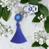 Blue evil eye keychain with tassel on a marble surface with white flowers and green leaves.