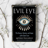 Book cover of 'The Evil Eye' on a marble surface with decorative branches.