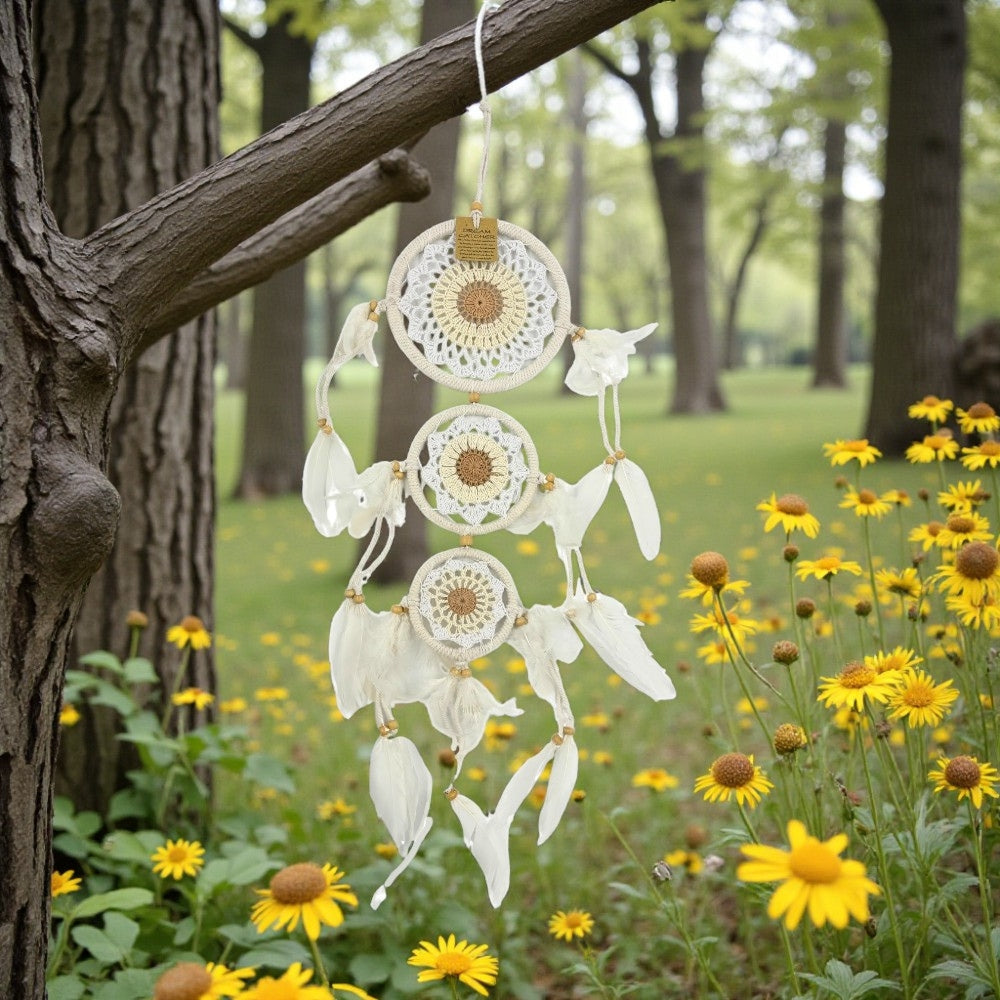 White dreamcatcher hanging from a tree branch in a forest with yellow flowers.