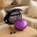 A purple steel tongue drum and its colorful geometric patterned carrying bag are displayed on a wooden table in a cozy living room setting.