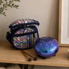 A small patterned fabric bag with a blue strap sits on a wooden shelf beside a metallic blue and purple steel drum, with two drum mallets in front. A beige wall, framed art, and a plant branch are visible in the background.