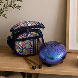 A small patterned fabric bag with a blue strap sits on a wooden shelf beside a metallic blue and purple steel drum, with two drum mallets in front. A beige wall, framed art, and a plant branch are visible in the background.