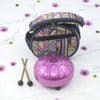 Purple metal tongue drum with patterned carrying bag and mallets on a white background with flowers.