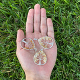 Three silver tree-of-life pendant charms made from amber-colored stone are displayed on an open hand, set against a background of green grass.