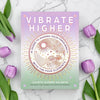 Book cover for 'Vibrate Higher' by Julieta Suárez Valente, styled in a flat-lay on a white marble surface with purple tulips. The cover features a pastel gradient transitioning from lavender to mint green, with a central circular illustration of suns, stars, and elements in gold and pink.