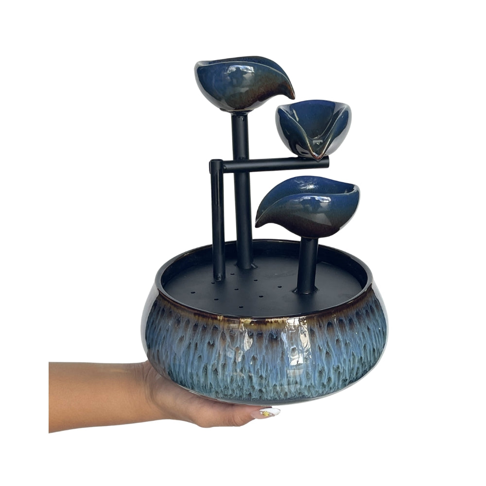 A three-tiered ceramic waterfall fountain with a textured blue and black glazed finish, held by a person's hand against a plain white background.