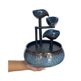 A three-tiered ceramic waterfall fountain with a textured blue and black glazed finish, held by a person's hand against a plain white background.