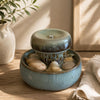 A small ceramic tabletop waterfall fountain with a light blue glazed base, a darker blue-green top tier, and smooth stones inside, styled on a wooden surface with a vase and cloth nearby.