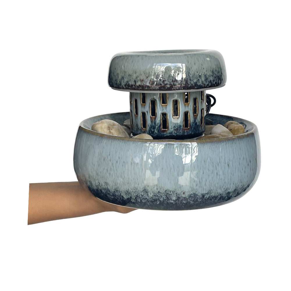 A ceramic two-tiered tabletop fountain with a blue and white glaze, held by a hand against a plain white background.